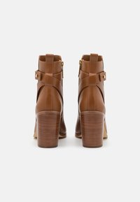 Botas de tobillo de cuero marrón con un tacón de madera apilada, detalle de correa decorativa y cierre de cremallera lateral. Textura suave y diseño elegante.