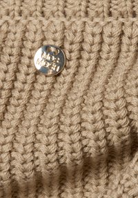 Beżowa dzianina z wyraźnym prążkowaniem, z okrągłą srebrną metką z wytłoczonym napisem "Moshi". Tekstura wygląda na grubą i przytulną.