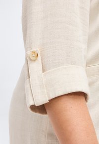 Τυλιγμένο μανίκι από μπεζ λινό ύφασμα, στερεωμένο με μια ταμπ που ταιριάζει από το ίδιο ύφασμα και ένα μπεζ κουμπί πάνω από ένα γυμνό χέρι.