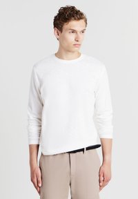 Giovane uomo che indossa una camicia bianca a maniche lunghe con trama, infilata in pantaloni beige con una cintura nera, mentre guarda di lato su uno sfondo neutro.