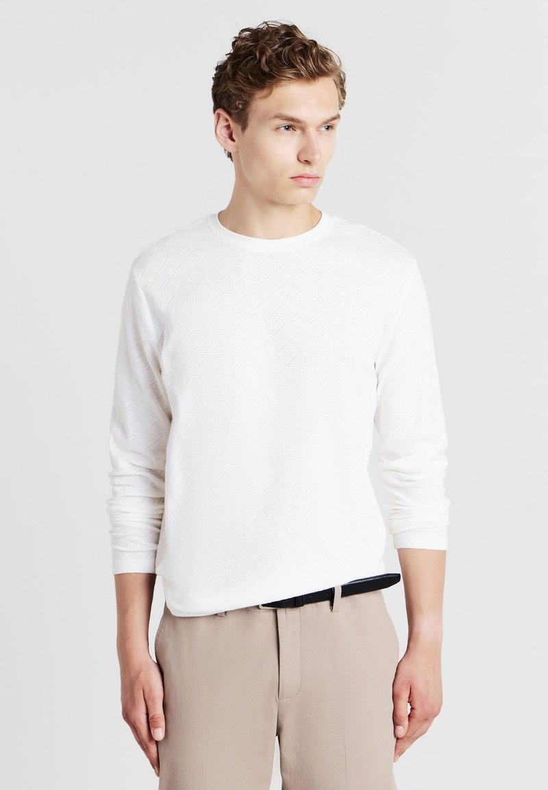 Giovane uomo che indossa una camicia bianca a maniche lunghe con trama, infilata in pantaloni beige con una cintura nera, mentre guarda di lato su uno sfondo neutro.