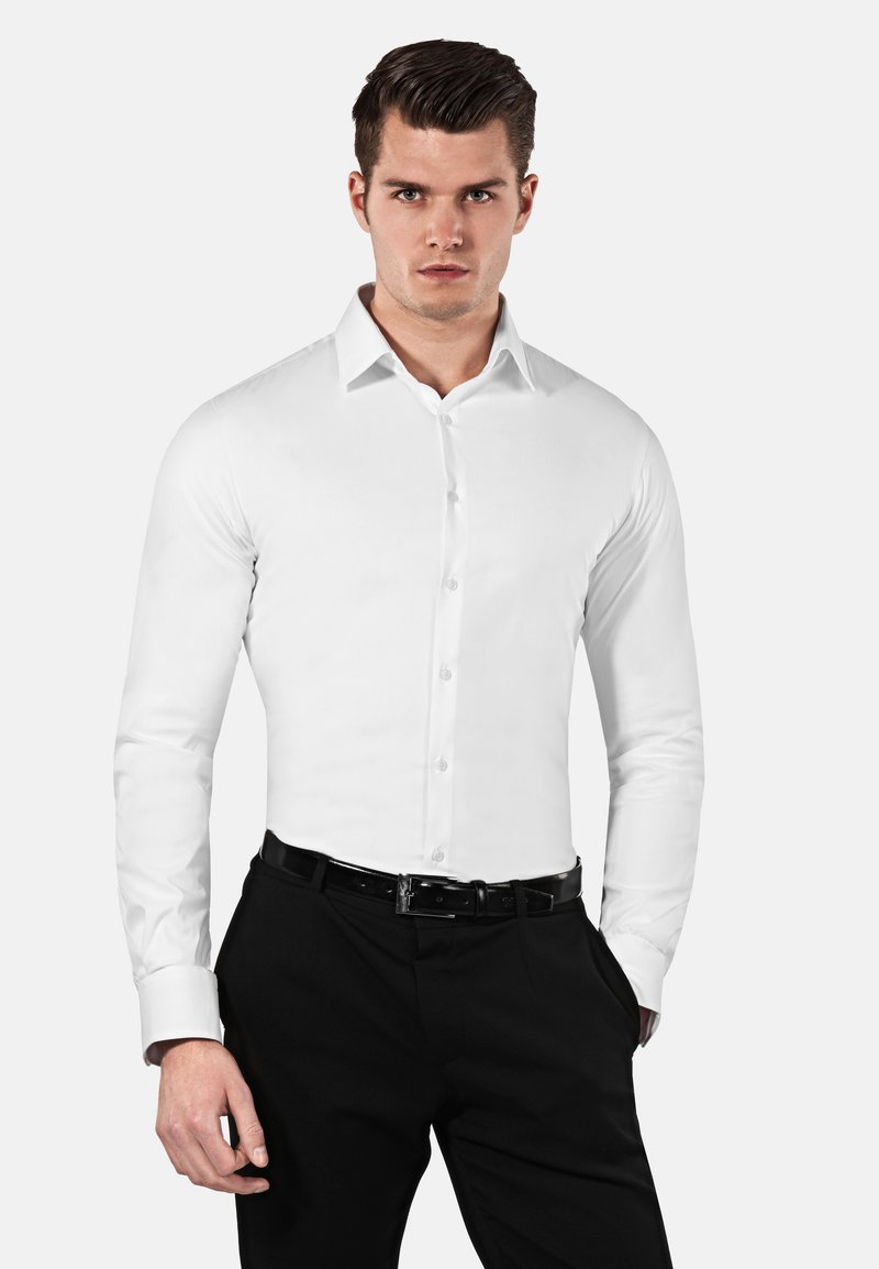 Chemise blanche à manches longues et boutonnière, en tissu lisse, avec un col pointu et une coupe ajustée, associée à un pantalon noir et une ceinture.