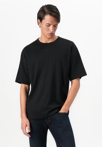 Fiatal férfi lefelé néz, sima fekete, oversize pólóban és sötétkék farmernadrágban, egyik kezével zsebében, fehér háttér előtt.