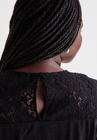 Top en dentelle noire avec une fermeture au dos en forme de goutte d'eau et un bouton, ainsi qu'un panneau en tissu uni contrastant. Texture lisse et motif floral en dentelle.