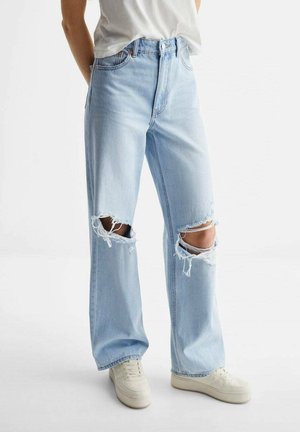 Persona vistiendo vaqueros rasgados de color azul claro con desgarrones en ambas rodillas, zapatillas blancas y una camiseta blanca de manga corta metida por dentro.