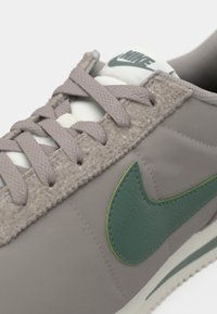 Pelēki sporta apavi ar teksturētu virsmu, zaļu Nike logo, gaiši beigu akcentiem, plakaniem šņorēm un gumijas zoli ar nelielu protektora rakstu.