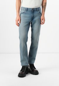 Een persoon staat gekleed in lichtblauwe jeans en zwarte stevige boots, met een tattoo zichtbaar op hun linkerarm, tegen een eenvoudige witte achtergrond.