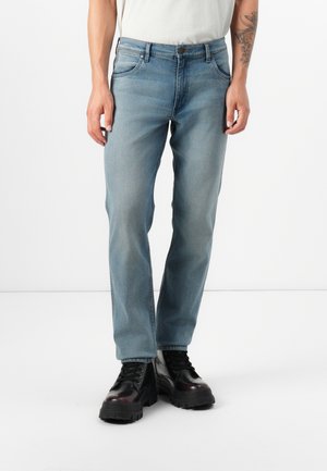 Una persona está de pie vistiendo jeans azul claro y botas negras gruesas, con un tatuaje visible en su brazo izquierdo, contra un fondo blanco liso.
