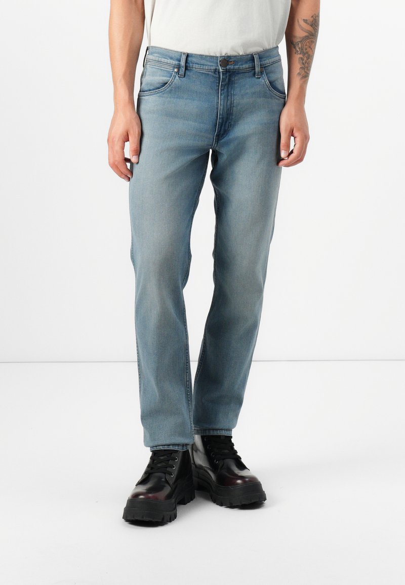Een persoon staat gekleed in lichtblauwe jeans en zwarte stevige boots, met een tattoo zichtbaar op hun linkerarm, tegen een eenvoudige witte achtergrond.