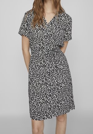 Femme portant une robe noire à manches courtes avec un motif floral blanc et une ceinture nouée, debout devant un fond gris uni.