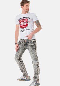 Weißes T-Shirt mit rographischem und schwarzem Druck, graue verwaschene Jeans mit mehreren Nähten und graublaues Sneaker mit Farbakzenten.