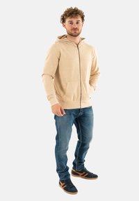 Felpa con zip beige, dotata di tasche frontali, in materiale di cotone e con logo. Indossata con jeans in denim blu e sneakers nere con accenti arancioni.