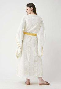 White textured maxi dress with square patterns, featuring wide sleeves and a yellow belt. Worn with brown flat sandals.