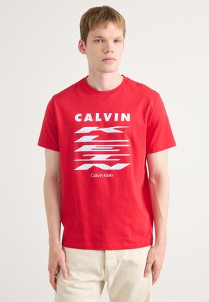 Červené bavlněné tričko s výrazným bílým grafickým textem "CALVIN" a stylizovaným "Klein" s diagonálními liniemi, kulatým výstřihem a krátkými rukávy.