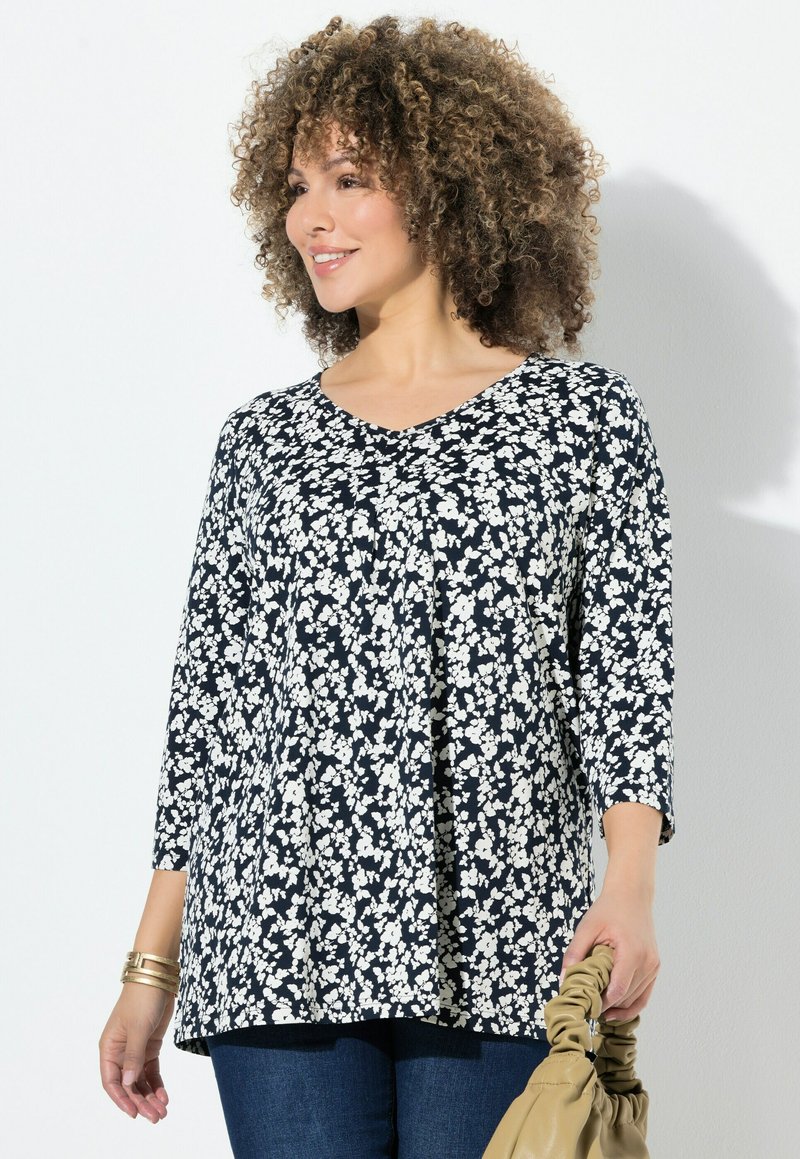 Námořnická modrá blůza s bílým květinovým vzorem, výstřih do V a rukávy tři čtvrtě. Lehce padnoucí látka volně splývá. Drží béžovou kabelku.