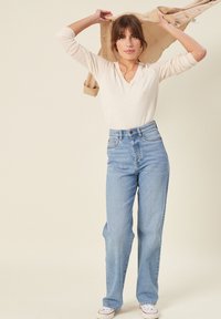 Jasno beżowa bluzka z długim rękawem, jasnoniebieskie jeansy z wysokim stanem i białe sneakersy. Modelka trzyma beżową kurtkę nad głową.