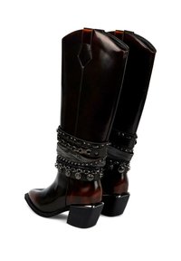 Cizme cowboy din piele maro, cu toc gros, decorate cu elemente metalice detaliate și curele ornamentale în jurul gleznei.