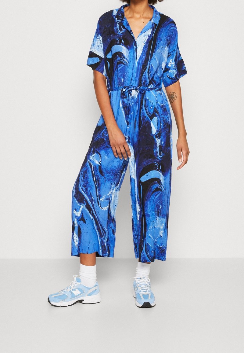 Personne portant une combinaison bleu marbré à manches courtes, taille cintrée, associée à des chaussettes blanches et des baskets bleu clair.