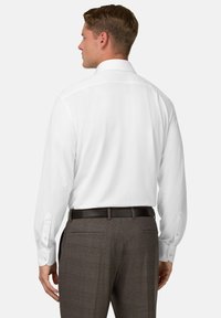 Uomo che indossa una camicia bianca a maniche lunghe infilata in pantaloni a quadri marroni con una cintura nera, visto da dietro.