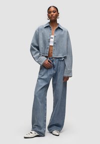 Giacca di denim azzurro chiaro cropped con grandi bottoni e maniche larghe, abbinata a jeans azzurri chiari a vestibilità ampia e scarpe bianche.