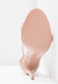 Nude lakované vysoké podpatky s úzkým stiletto a texturovanou podrážkou; obsahují tenký pásek kolem kotníku pro podporu.