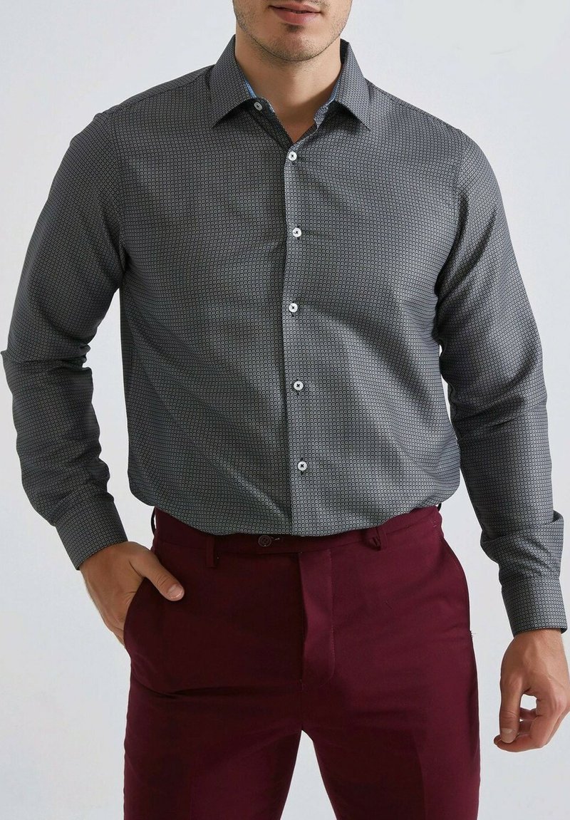Chemise grise à motifs avec un col boutonné, présentant un design géométrique et des poignets contrastants, portée avec un pantalon bordeaux.