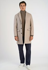 Abrigo de lana beige claro con solapas, seis botones y bolsillos frontales, combinado con un jersey de cuello alto marrón y pantalones azul marino, junto a zapatillas blancas.