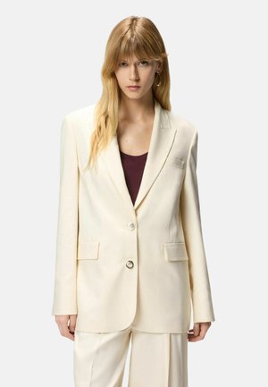 Giovane donna con lunghi capelli biondi indossa un blazer crema oversize e pantaloni coordinati sopra una maglia scura, in piedi davanti a uno sfondo semplice.