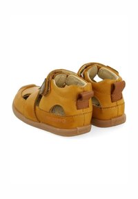 Sandalias de cuero marrón con talón cerrado, lados abiertos y correa ajustable de Velcro. Suela de goma texturizada y acentos cosidos.