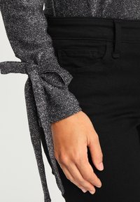 Handen vilar vid höften, iklädd svarta byxor och en glittrig silverfärgad långärmad topp med en knuten rosett vid handleden.