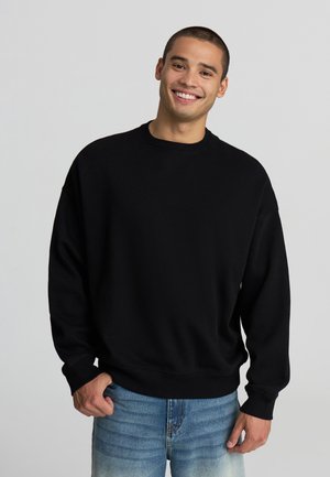 Felpa nera oversize in misto cotone, con collo rotondo, polsini a coste e vestibilità comoda, abbinata a jeans azzurri chiari.