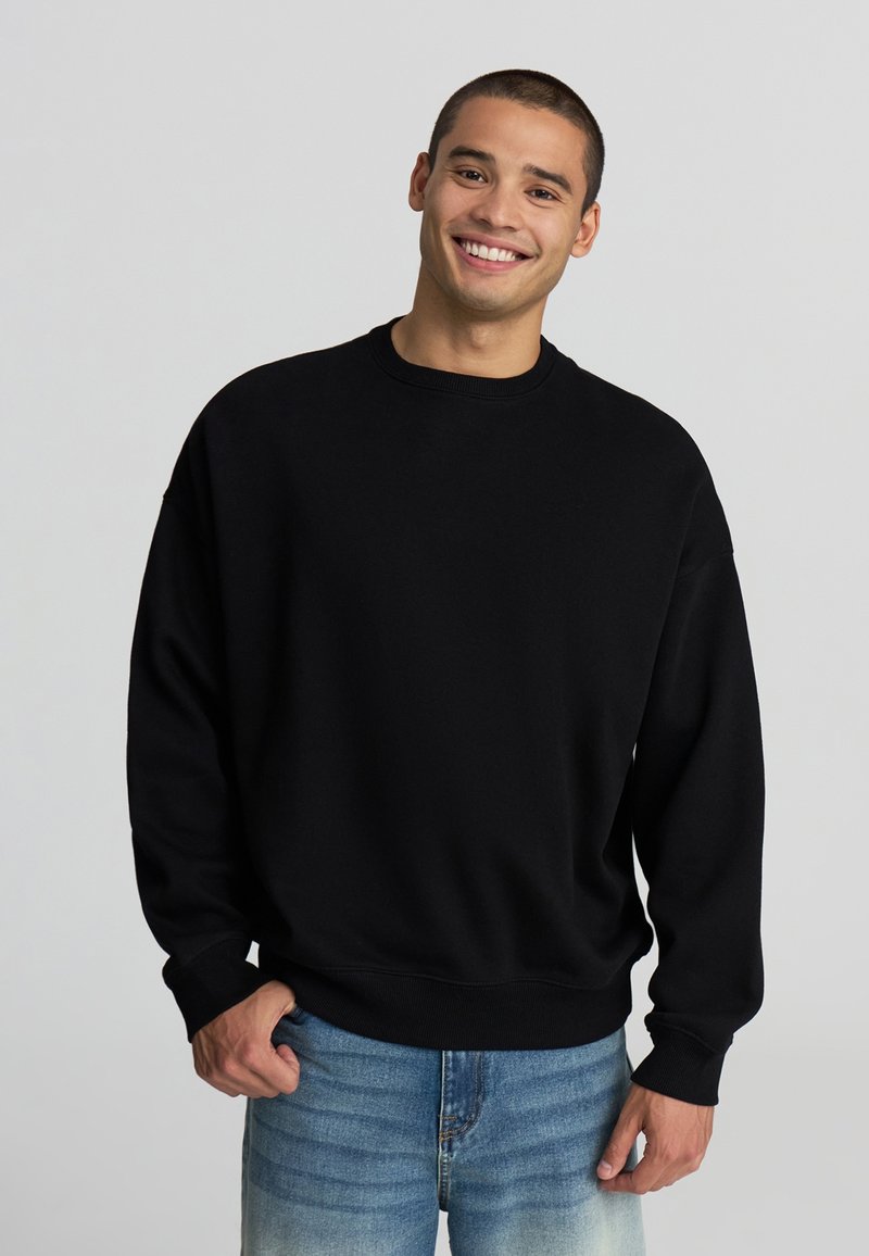 Felpa nera oversize in misto cotone, con collo rotondo, polsini a coste e vestibilità comoda, abbinata a jeans azzurri chiari.