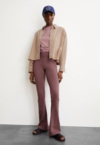 Light beige button-up shirt over a mauve fitted top and flared mauve pants, paired with black slide sandals and a dark blue cap.