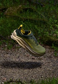Chaussure de trail running verte avec un dessus en mesh texturé, des accents jaunes, une semelle rembourrée proéminente et des perforations pour la respirabilité.