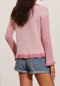 Femme aux longs cheveux noirs portant un pull à manches longues rose à motifs avec un ourlet volanté et un short en denim bleu retroussé, de dos.
