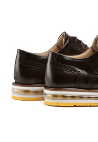 Braune Lederschuhe mit Brogue-Verzierungen, runder Zehenpartie und einer gepolsterten, transparenten Sohle. Gelber Akzent am Sohlenrand.