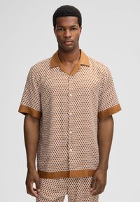 Man in een bruin en wit gestreept kort overhemd met bijpassende broek, staande tegen een effen achtergrond.