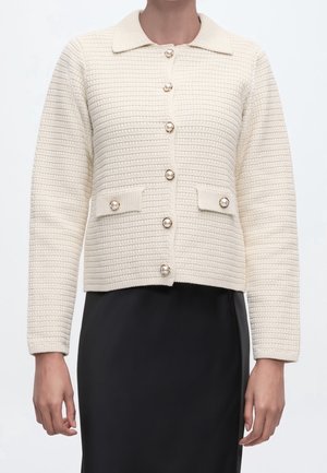 Femme portant un cardigan en maille texturée crème avec des boutons en perle et des poches à rabat sur une jupe noire.