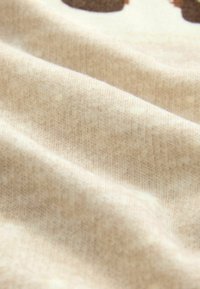 Weicher beige Stoff mit feiner gerippter Struktur und sanften Falten, Teil eines größeren Textilartikels mit dunklen braunen Druckelementen.