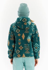 Hoodie in pile verde acqua, con cappuccio, caratterizzato da motivi geometrici in marrone chiaro e acqua. Abbinato a pantaloni blu chiaro e a un berretto coordinato.