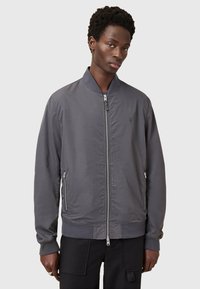 Giacca bomber grigia con zip frontale, colletto e polsini a costine, due tasche laterali con zip e un logo discreto sul petto. Realizzata in tessuto morbido.