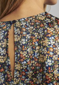 Top con motivo floreale in tessuto trasparente blu scuro, caratterizzato da piccoli fiori arancioni, bianchi e verdi. Apertura a chiave sul retro con chiusura a bottone nera.