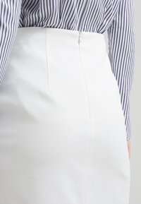 Bílá texturovaná sukně se zapínáním na zip na zadní straně, zkombinovaná s košilí na knoflíky v námořnické modré a bílé pruhy. Má přiléhavý střih a jemné švy.