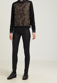 Černá košile s dlouhým rukávem, která má přední stranu s kamuflážovým vzorem, zkombinovaná s černými úzkými džíny a černými botami se cvočky.