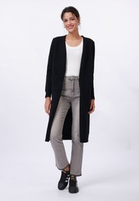 Long black cardigan with open front and two front pockets, worn over a white top and gray jeans. Black sneakers add contrast to the outfit.