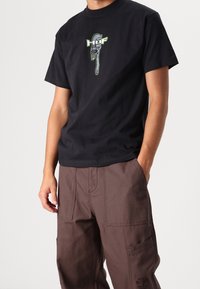 T-shirt en coton noir avec un graphique d'une clé à molette en vert et blanc. Associé à un pantalon cargo marron ample avec poches.