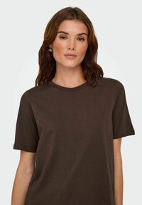 Donna con lunghi capelli castani che indossa orecchini d'argento e una maglietta a maniche corte color marrone scuro, rivolta in avanti con un'espressione neutra.