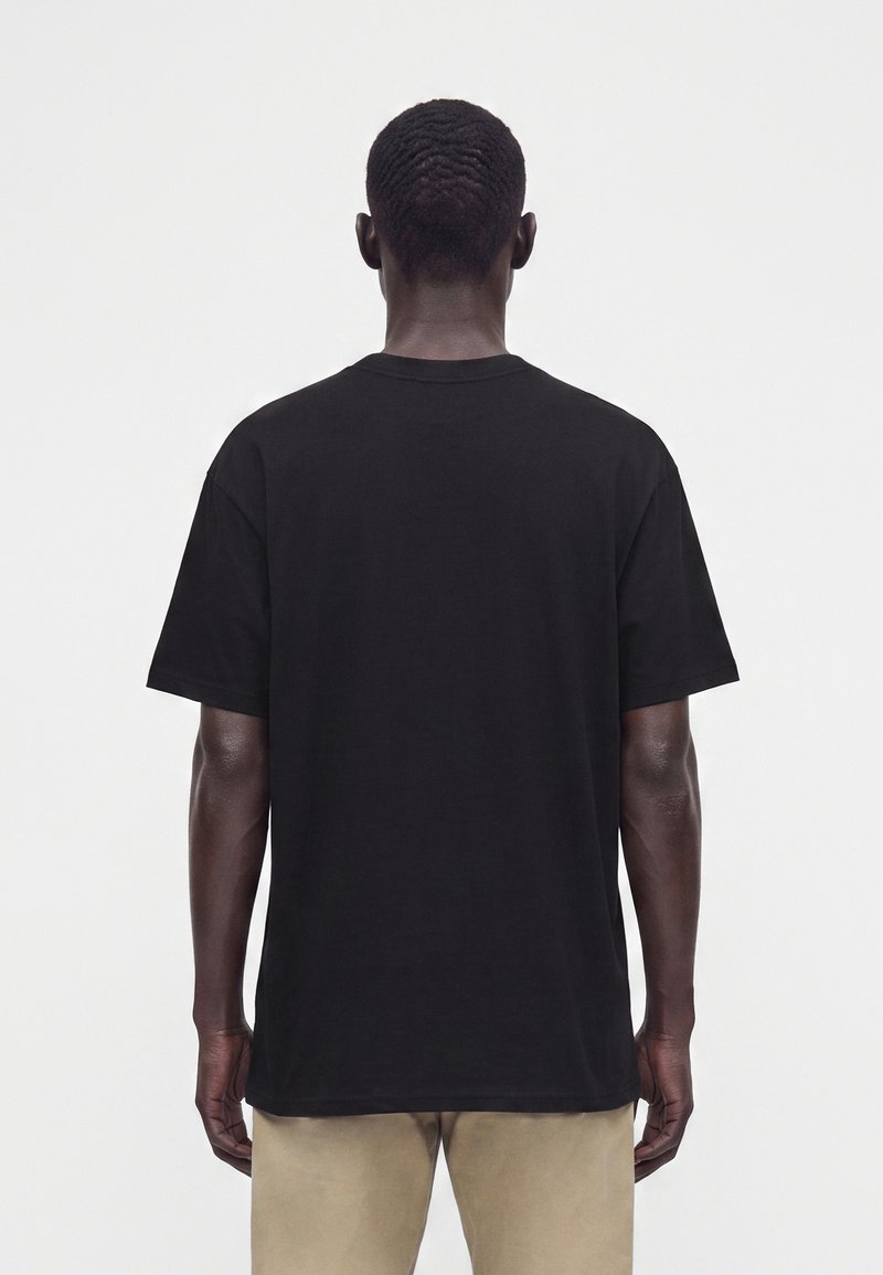 Uomo con capelli corti visto di spalle, che indossa una t-shirt nera semplice a maniche corte e pantaloni beige su sfondo bianco.