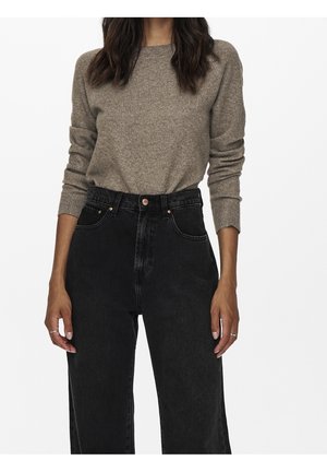 Persoon die een beige trui met lange mouwen draagt, ingestopt in zwarte high-waist jeans, met handen langs het lichaam.