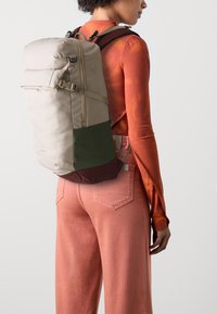 Beige reppu, jossa on vihreitä ja burgundinpunaisia yksityiskohtia, varustettu rullatoppisuunnittelulla, sivuhihnalla ja pehmustetuilla olkahihnoilla.